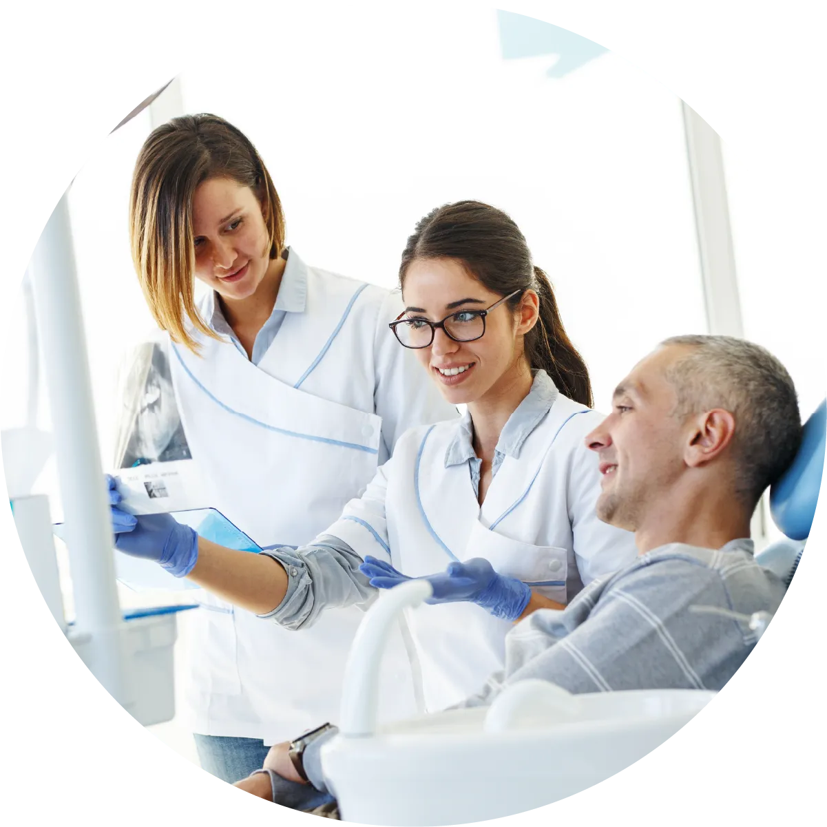 Two female dental professionals in scrubs are reviewing what appears to be an X-ray with a male patient in a dental chair. 