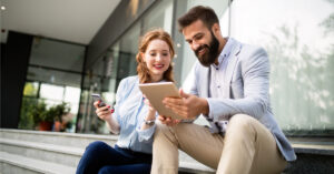 Smiling man and woman sitting outdoors, looking at a tablet together while holding a smartphone and chatting.