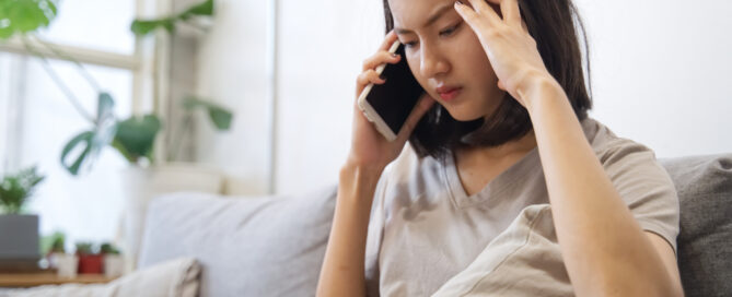 Person sitting on a couch at home, talking on a smartphone and holding their temples as if stressed or overwhelmed.
