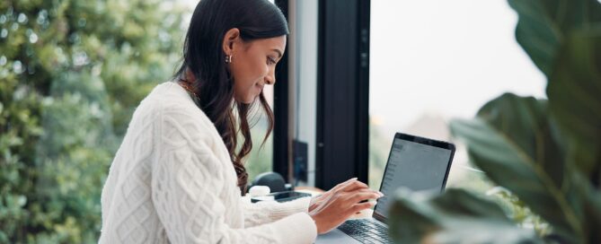 Woman in a white sweater using a laptop at a desk by a large window with greenery outside