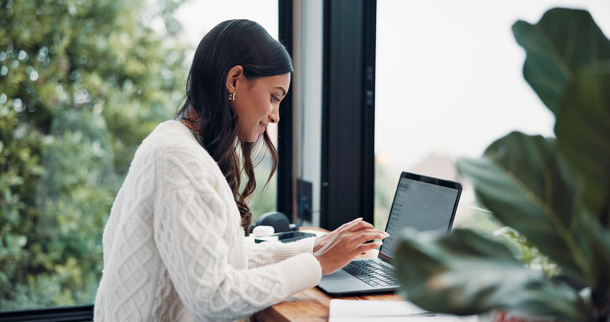 Woman in a white sweater using a laptop at a desk by a large window with greenery outside