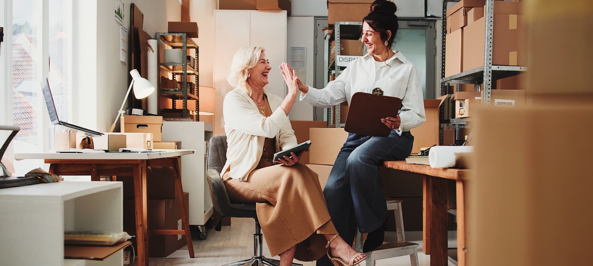 Two female coworkers sitting at a desk and high-fiving each other while smiling.