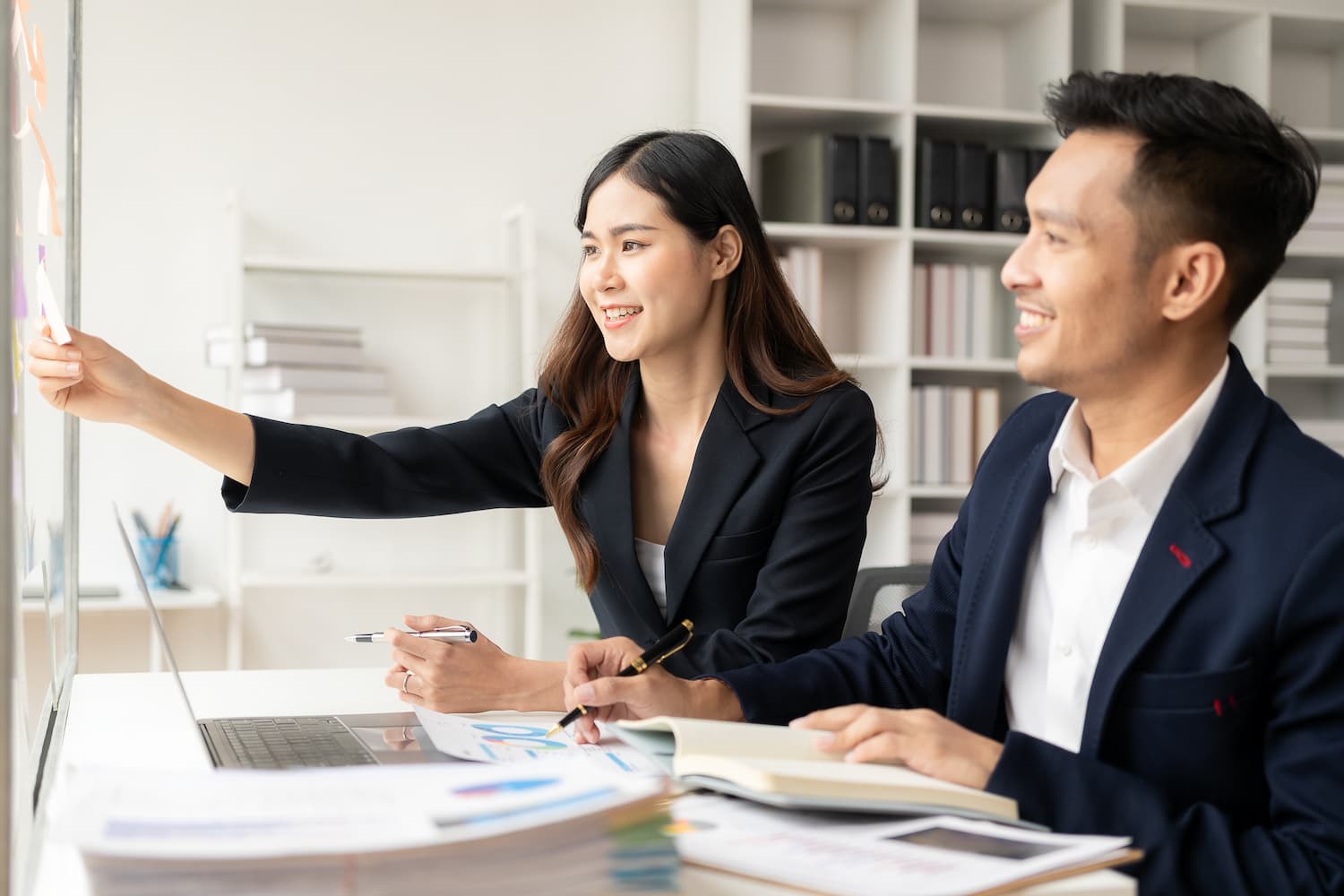 Two coworkers reviewing notes and placing sticky notes on a glass board during a planning session.