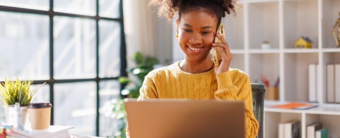 A young woman in a yellow sweater smiles while talking on the phone and working on a laptop at a desk in a bright home office.