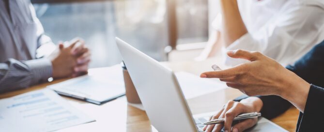 Two professionals meeting at a table with a laptop, documents, and a tablet during a workplace discussion.