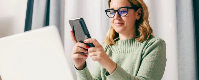 Woman in a green sweater and black glasses looking at a phone in front of an open laptop