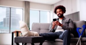 A man with a leg cast and crutches sits on a couch while using a smartphone.