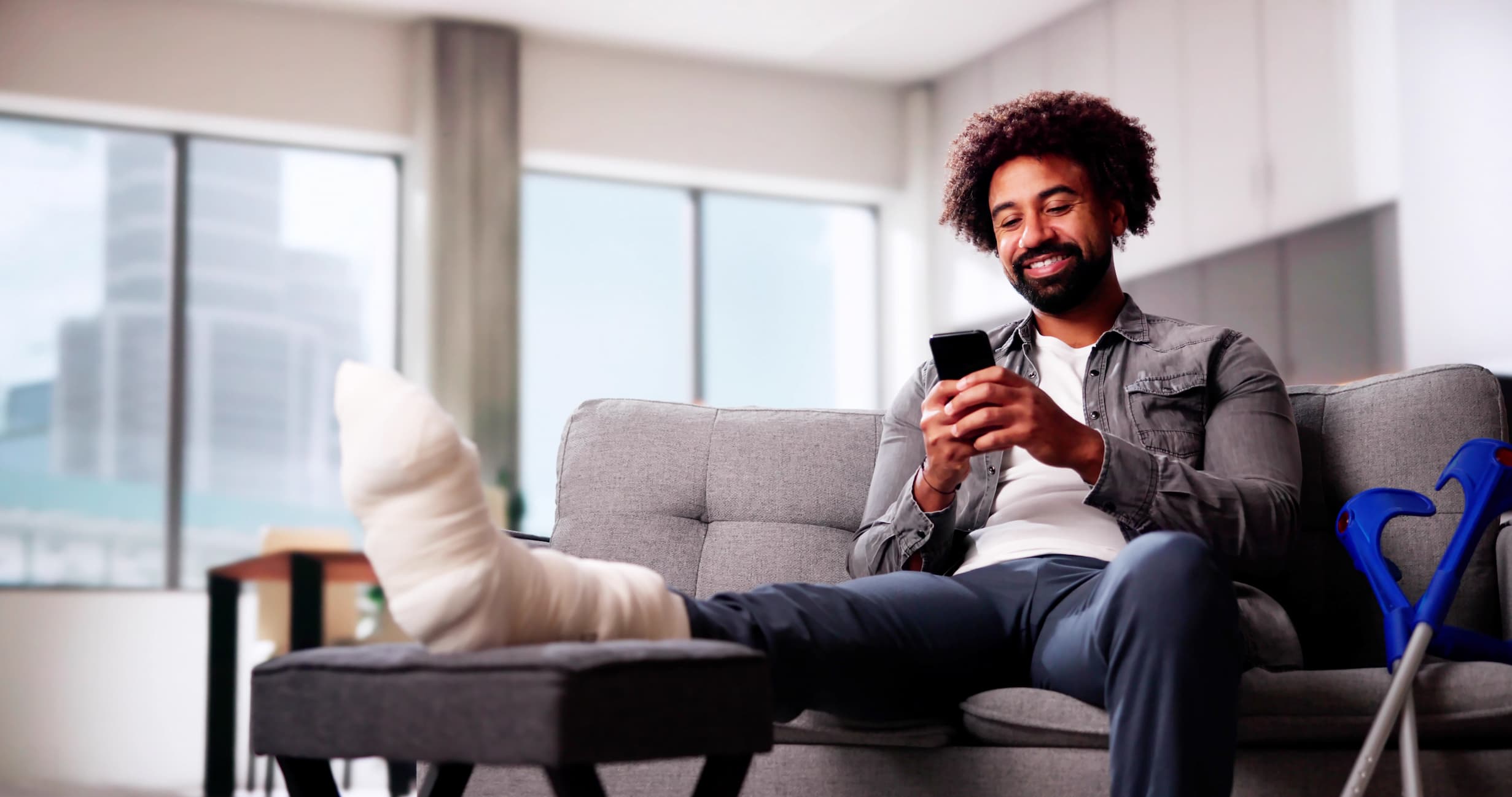 AdobeStock_1026442515 A man with a leg cast and crutches sits on a couch while using a smartphone.