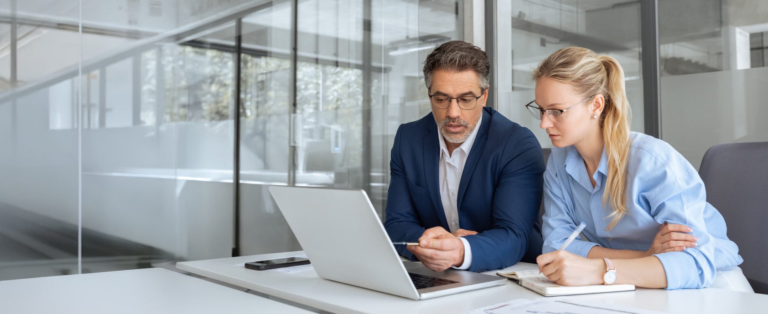 AdobeStock_1719468947 A man in a blue suit and a woman in a light blue shirt sit at a desk in a modern office, looking at a laptop screen together.