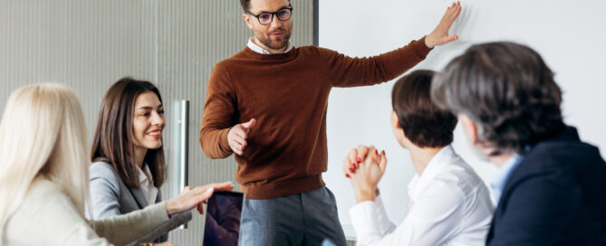 Business professional presenting to a small group during a meeting while colleagues listen around a conference table.