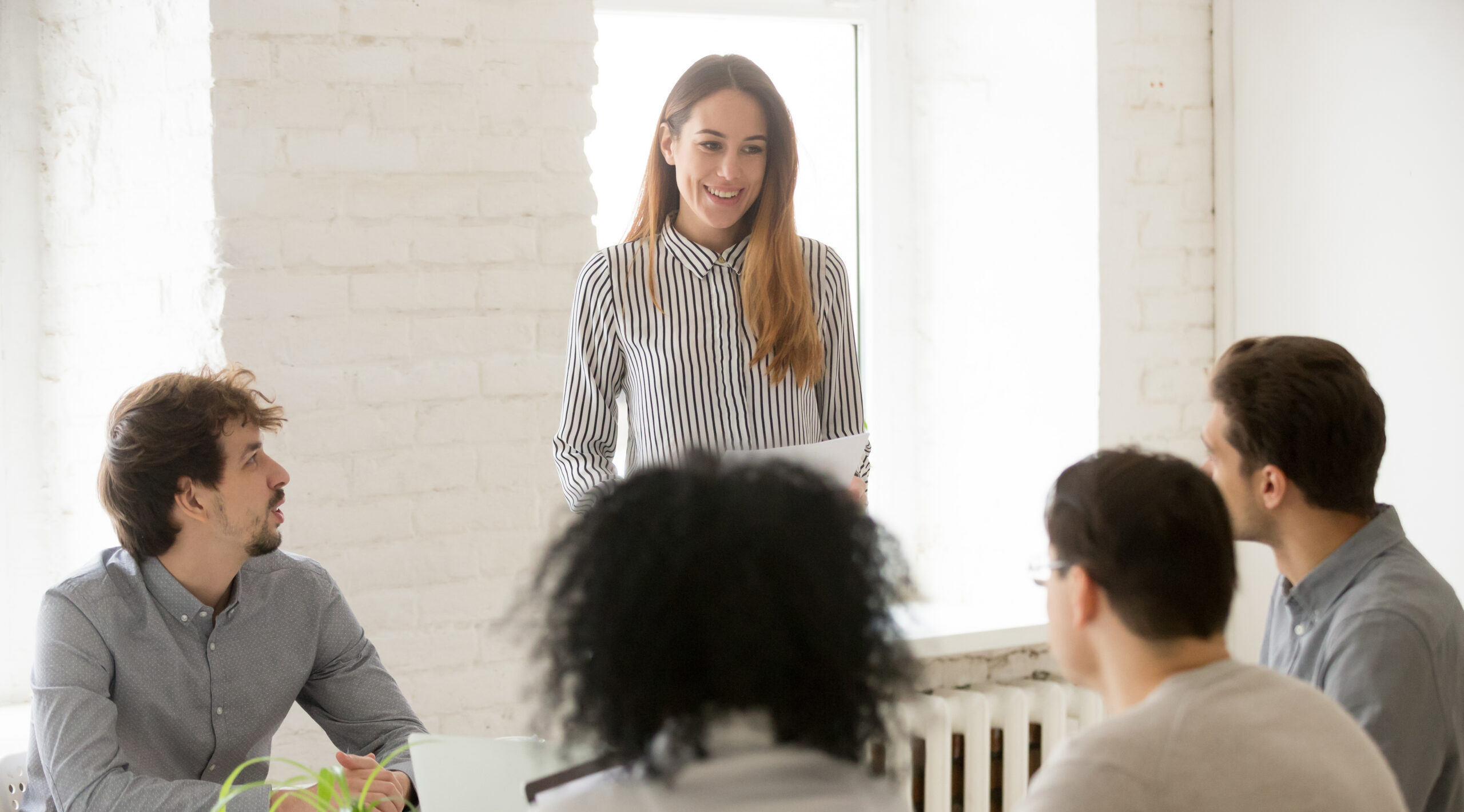 Smiling woman standing at the head of a conference table, leading a meeting with four seated colleagues in a bright office.