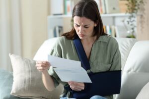 A woman with her arm in a sling sits on a sofa, looking worried while reading a bill.