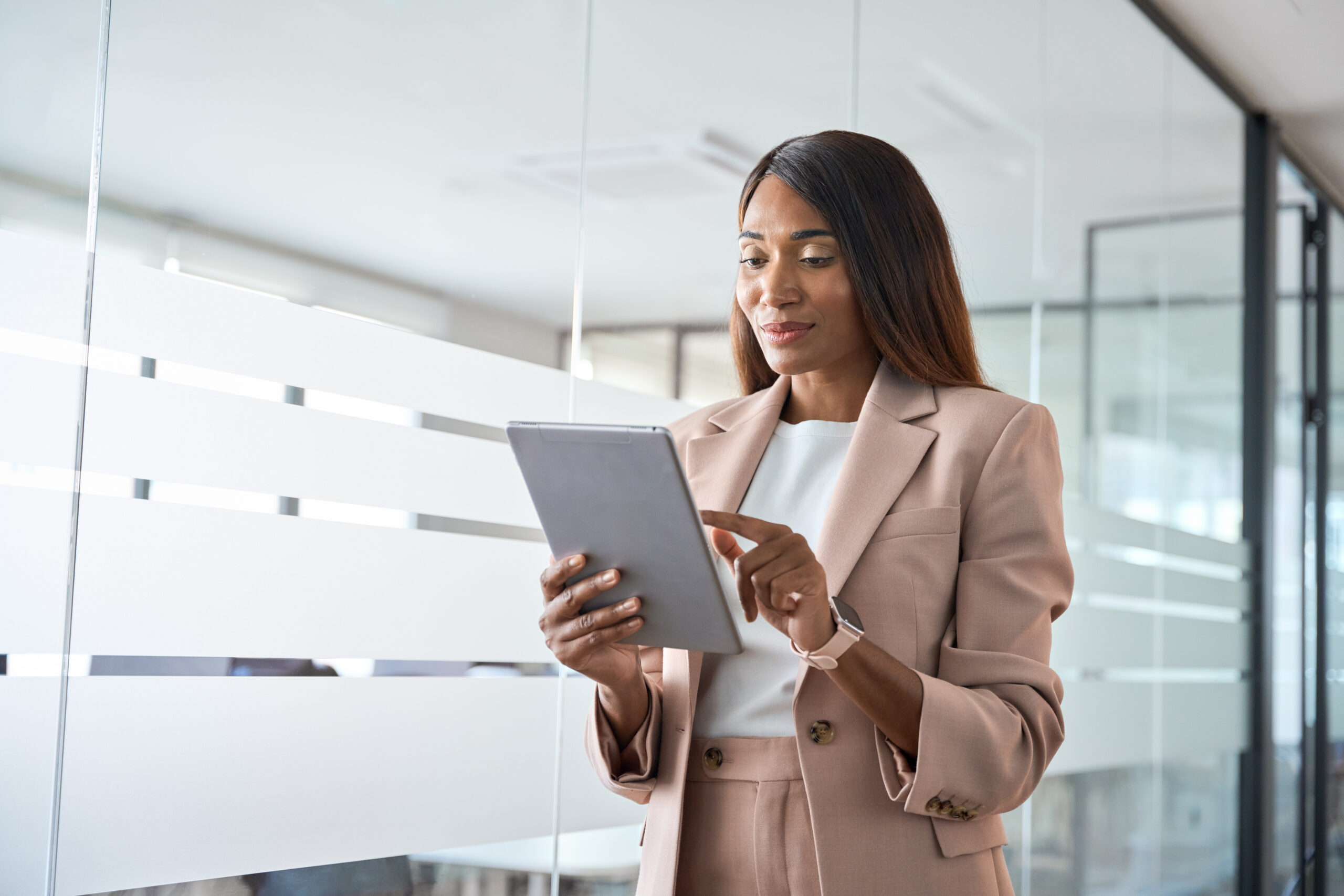 Businesswoman in a beige suit using a tablet in a bright, modern office with glass walls.