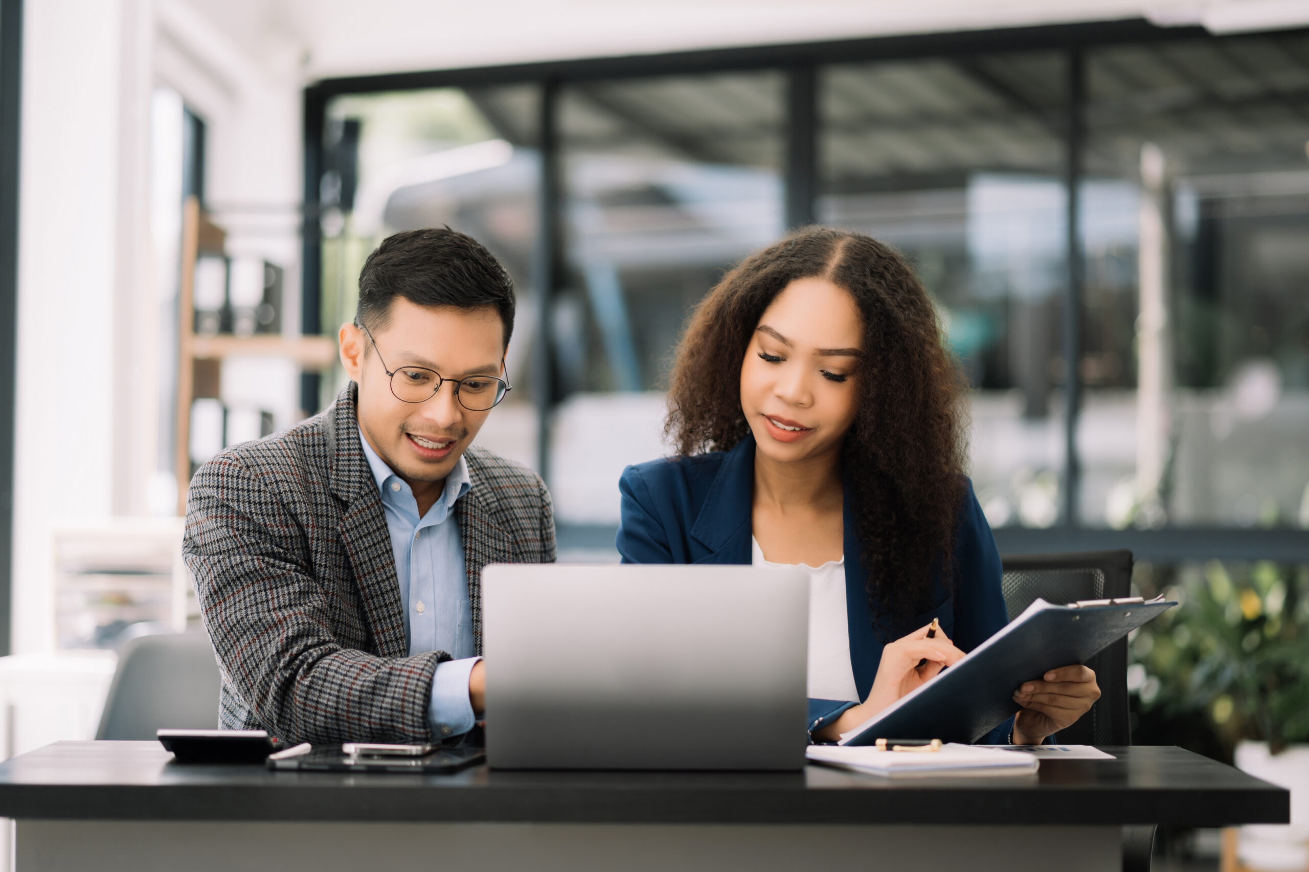 Man in a light blue shirt holding a coffee cup and woman in a black blazer reviewing a document together at a desk with a laptop, in a bright office with large windows.